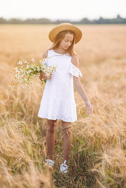 Happy child in wheat field. Beautiful girl in white dress in a straw hat with ripe wheat in hands - Foto, Imagem