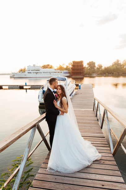 Schönes luxuriöses Paar, der Bräutigam im schwarzen Anzug und die Braut mit Schleier und weißem Brautkleid, Händchen haltend, am Pool auf der Seebrücke neben der Jacht und dem Schiff - Foto, Bild