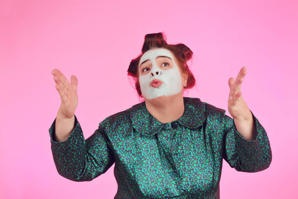 humorous portrait of overweight caucasian woman in bathrobe and curlers talking on pink isolated background - Photo, Image