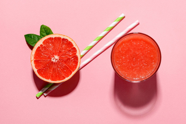Glass of freshly squeezed grapefruit juice with pulp, half grapefruit and two drinking straws  cast shadows in bright light on a pink background. Vegetarian, raw food diet and healthy eating. Top view. - Foto, immagini