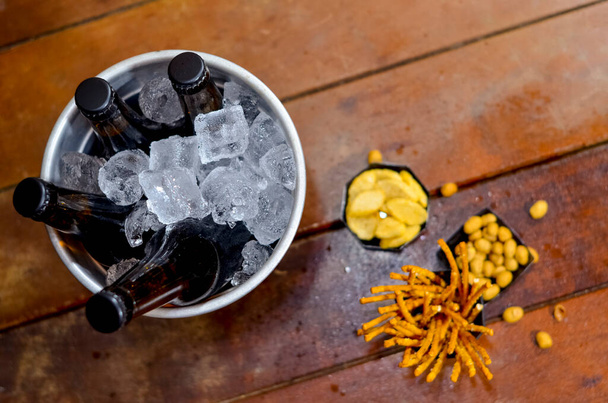 Черное ведро со льдом с четырьмя бутылками пива внутри на деревянном столе и с некоторыми вкусными закусками. - Фото, изображение