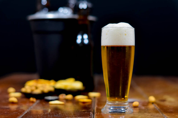 A glass full of beer in the foreground with a bottle of beer to the side and a black bucket with beers inside, some savory snacks on a wooden table with a black background. - Photo, Image