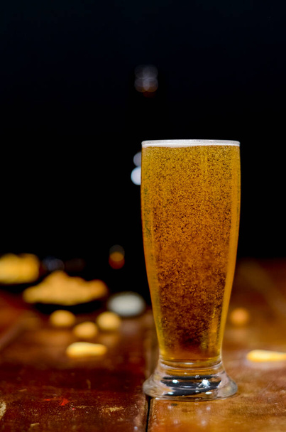 Ένα ποτήρι γεμάτο μπύρα στο μπροστινό μέρος με ένα μπουκάλι μπύρα στο πλάι και ένα μαύρο κουβά με μπύρες μέσα, μερικά αλμυρά σνακ σε ένα ξύλινο τραπέζι με μαύρο φόντο. - Φωτογραφία, εικόνα