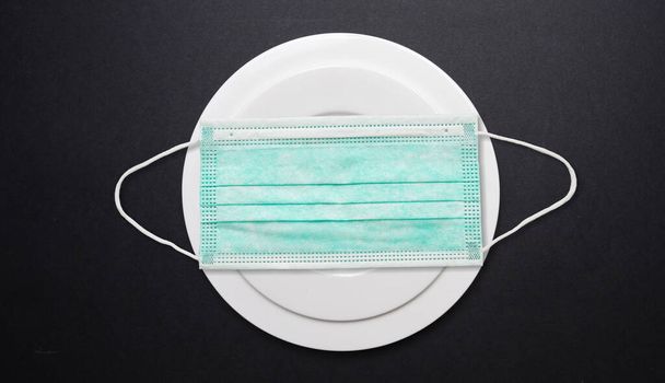 Coronavirus für Essen und Restaurantkonzept. Gesichtsschutzmaske auf einem Teller vor schwarzem Hintergrund, Kopierraum. COVID verbreitete Präventionsmaßnahme - Foto, Bild