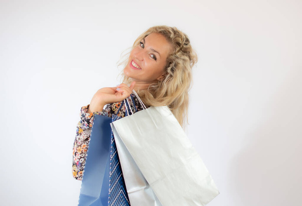 Çiçekli elbiseli güzel sarışın kadın beyaz arka planda alışveriş torbalarıyla el kol geziyor. - Fotoğraf, Görsel