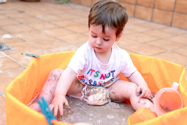 Dziecko bawiące się piaskiem w pomarańczowym plastikowym basenie. Na tarasie domu. - Zdjęcie, obraz
