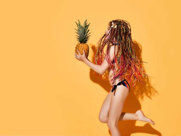 Side shot of girl with colorful afro bunches jumping on one leg in bikini holding pineapple. Three quarter shot isolated on orange, copy space - Photo, image