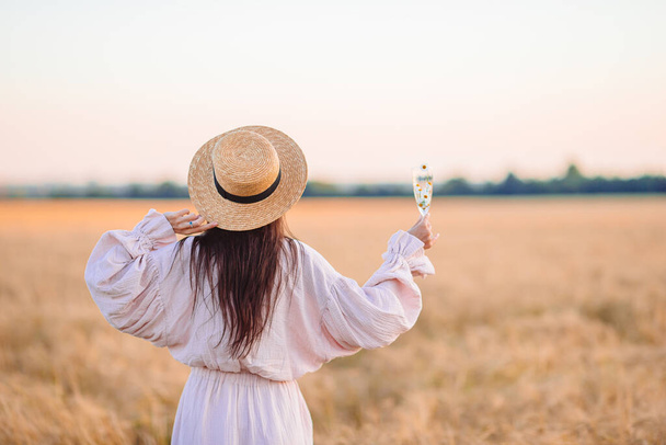 Vue arrière de la fille dans le champ de blé. Belle femme en robe dans un chapeau de paille avec du blé mûr dans les mains - Photo, image