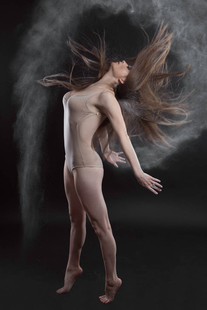 Latinská mladá žena s tenkou bílou pletí, stojící na špičkách pohybující se její dlouhé vlasy, které je s mastkem prášek, na sobě krémově zbarvené bodysuit, ve studiu s černým pozadím - Fotografie, Obrázek