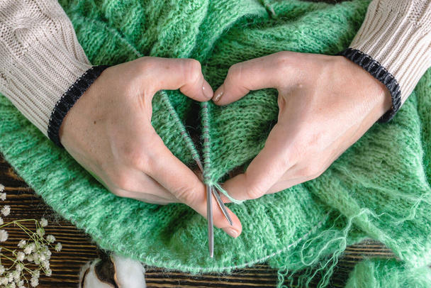 Warm cozy green sweater in the process of knitting and hands folded in the shape of a heart. Concept of your favorite hobby. - Фото, изображение