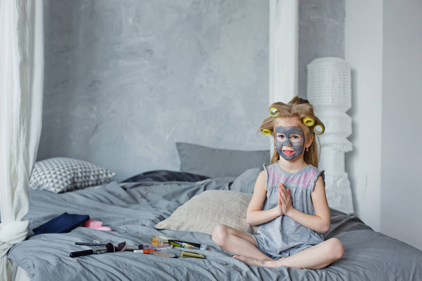 Uma menina feliz em rolos com uma máscara no rosto está sentada na cama no quarto, fazendo procedimentos cosméticos. Conceito de um salão de beleza, Spa e centro de Bem-Estar
. - Foto, Imagem