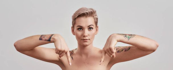 Look here. Portrait of half naked tattooed woman with short hair looking at camera pointing index fingers at the bottom isolated over light background - Photo, Image