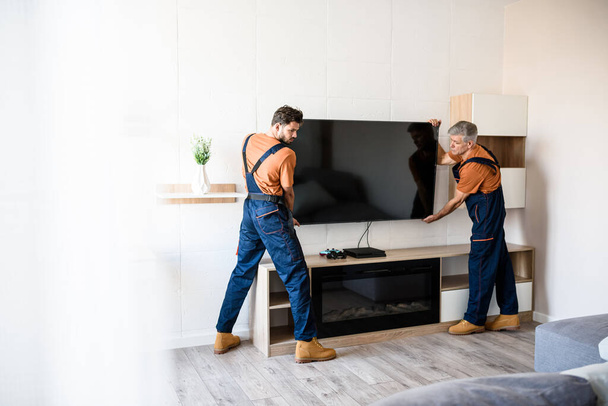 Delivered on time. Two handymen, workers in uniform hanging, installing tv television on the wall indoors. Repair and assembly service concept - Фото, зображення