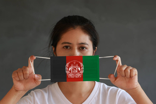 Μια γυναίκα με λευκό πουκάμισο με σημαία του Αφγανιστάν με υγιεινή μάσκα στο χέρι της και σήκωσε το μπροστινό πρόσωπο σε σκούρο γκρι φόντο. Μικροσκοπική προστασία μορίων ή ιών ή Covid 19. Έννοια της καταπολέμησης της ασθένειας. - Φωτογραφία, εικόνα