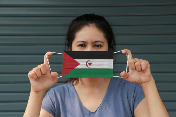 Eine Frau mit saharauisch-arabischer Flagge auf Hygienemaske in der Hand und hochgezogenem Gesicht auf dunkelgrünem Hintergrund. Winzige Partikel oder Viren Corona oder Covid 19 Schutz. Konzept zur Bekämpfung von Krankheiten. - Foto, Bild