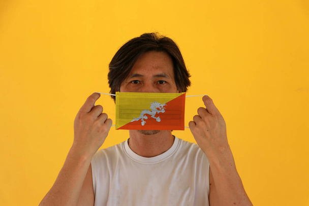 A man with Bhutan flag on hygienic mask in his hand and lifted up the front face on yellow background. Tiny Particle or virus corona or Covid 19 protection. Concept of Combating illness. - Photo, Image