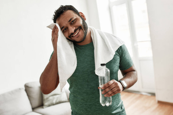 Tired but happy. Young man smiling at camera, holding water bottle and wiping sweat with towel while resting after training workout at home. Sport, healthy lifestyle - Fotoğraf, Görsel