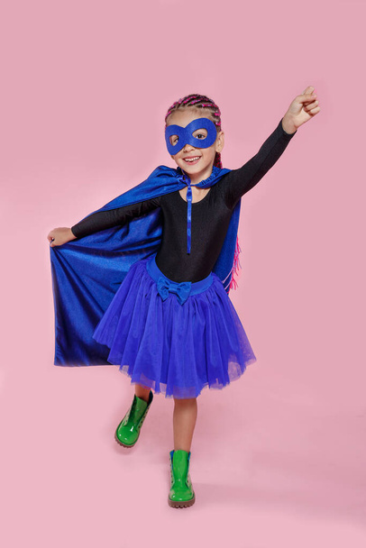 Close up portrait of a pretty little girl in superhero costume holding his hand and pointing up over pink background. - Photo, Image
