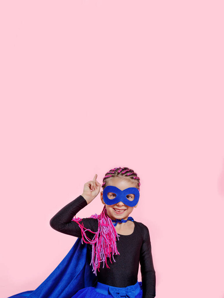 Lächelndes, entzückendes kleines Mädchen im Superheldenkostüm, das seine Hand hält und auf rosa Hintergrund zeigt. Raum für Text. - Foto, Bild