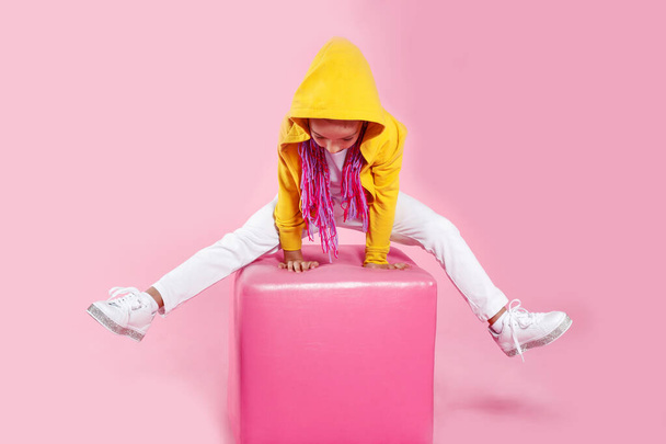 Hipster μικρό κορίτσι φορώντας λευκό τζιν, χρωματιστά ρούχα κάθεται με υψωμένα πόδια σε μια μαλακή καρέκλα στο στούντιο, πάνω από ροζ φόντο. Μοντέρνο παιδί.. - Φωτογραφία, εικόνα
