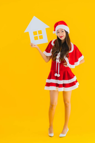 Portrait beautiful young asian woman wear christmas clothes and hat show home house sign with a lot of cash and money on yellow isolated background - Фото, зображення