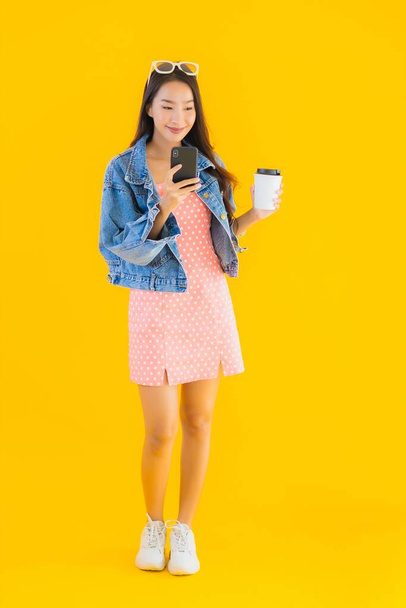 Πορτρέτο όμορφη νεαρή Ασιάτισσα γυναίκα με φλιτζάνι καφέ και έξυπνο κινητό τηλέφωνο σε κίτρινο απομονωμένο φόντο - Φωτογραφία, εικόνα