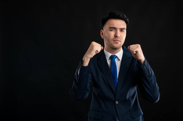 Portrait of business man wearing blue business suit and tie showing fists like fighting isolated on black background with copy space advertising area - Foto, Imagen