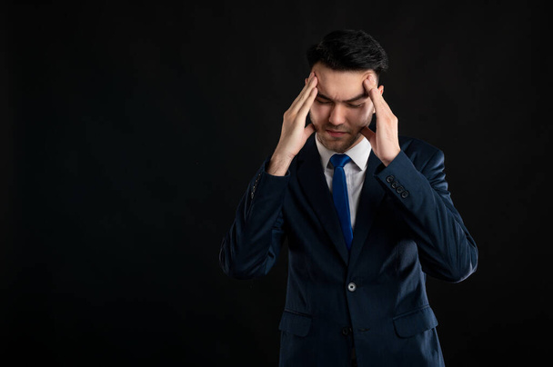 Portrait of business man wearing blue business suit and tie gesturing head ache isolated on black background with copy space advertising area - Photo, image