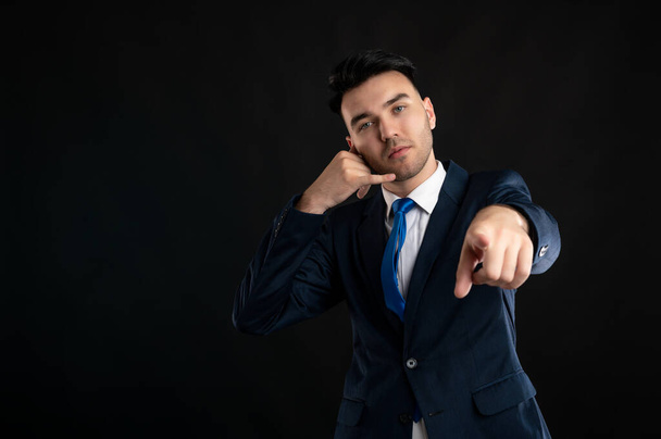 Retrato del hombre de negocios con traje de negocios azul y corbata que muestra el gesto de llamada o contacto apuntando a la cámara aislada sobre fondo negro con área de publicidad de espacio de copia - Foto, Imagen