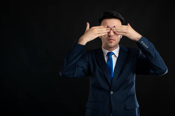 Portrait of business man wearing blue business suit and tie covering eyes like blind gesture isolated on black background with copy space advertising area - Foto, afbeelding