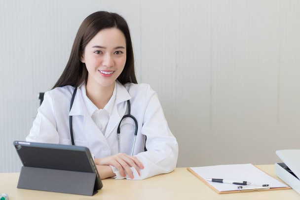 Aziatische mooie jonge glimlachende vrouwelijke arts zittend in het ziekenhuis. Op tafel heeft een klembord en een digitale tablet computer. - Foto, afbeelding