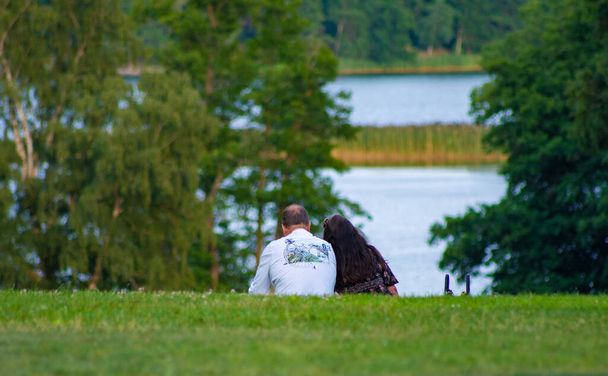 Um casal sentado em um gramado no fundo de um lago e árvores
 - Foto, Imagem