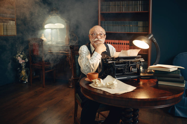Ηλικιωμένος συγγραφέας εργάζεται σε vintage γραφομηχανή στο γραφείο του. Ο γέρος με τα γυαλιά γράφει μυθιστόρημα λογοτεχνίας στο δωμάτιο με καπνό. - Φωτογραφία, εικόνα