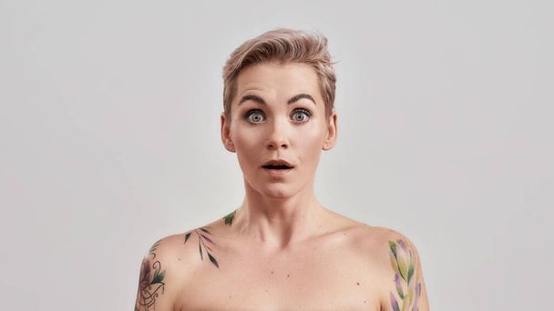 Shock. Portrait of a young attractive tattooed woman with pierced nose and short hair looking shocked or surprised at camera isolated over light background - Fotó, kép