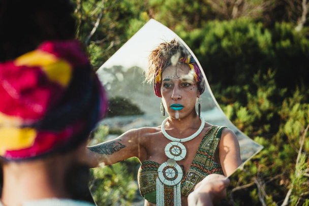 Frau in wilder Natur, mit afrikanischer Schminke und ethnischer Kleidung, die in den Spiegel schaut - Foto, Bild