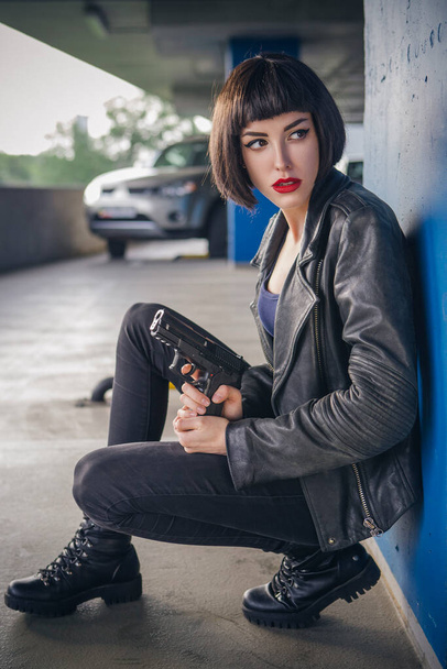 Linda morena sexy agente espião (assassino ou polícia) mulher em jaqueta de couro e jeans com uma arma na mão correndo atrás de alguém, para pegá-lo no estacionamento. - Foto, Imagem