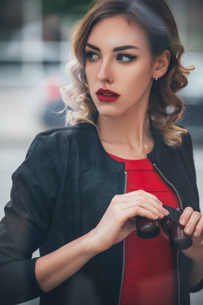 mooi brunette sexy spion agent (killer of politie) vrouw in lederen jas en rood luxe jurk met een verrekijker in haar hand kijken na iemand in europees stad. - Foto, afbeelding