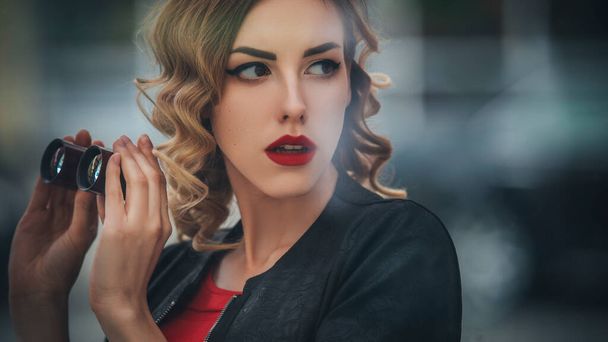 mooi brunette sexy spion agent (killer of politie) vrouw in lederen jas en rood luxe jurk met een verrekijker in haar hand kijken na iemand in europees stad. - Foto, afbeelding
