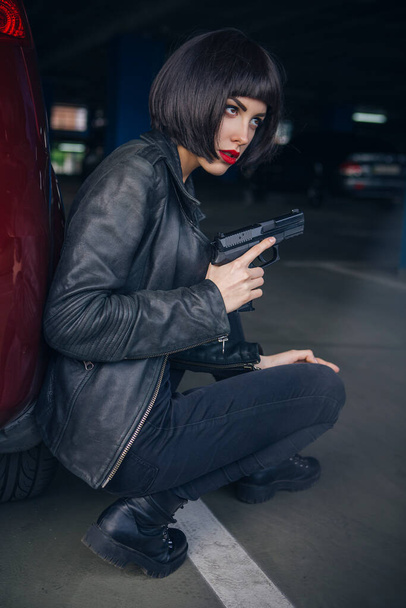 Красивая брюнетка сексуальный шпион агент (убийца или полиция) женщина в кожаной куртке и джинсах с пистолетом в руке бежит за кем-то, чтобы поймать его на парковке. - Фото, изображение