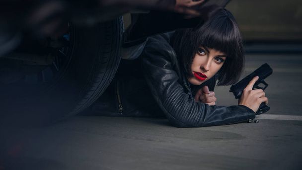 Beautiful brunette sexy spy agent (killer or police) woman in leather jacket and jeans with a gun in her hand running after someone, to catch him on parking. - Φωτογραφία, εικόνα