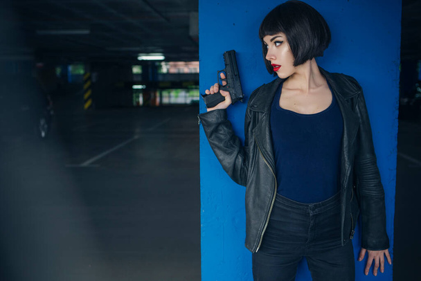 Bella bruna sexy agente spia (assassino o polizia) donna in giacca di pelle e jeans con una pistola in mano che corre dietro a qualcuno, per catturarlo nel parcheggio. - Foto, immagini