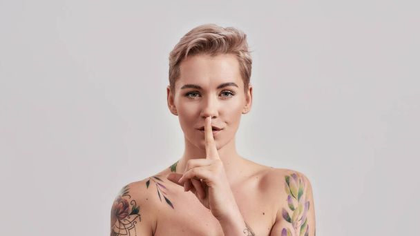 Keep silence. Portrait of half naked tattooed woman with short hair looking at camera, holding a finger on her lips isolated over light background - Photo, Image