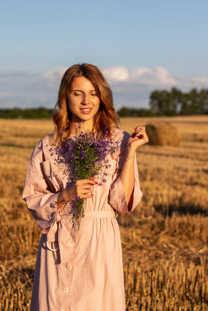 Beautiful young woman in vintage pink dress with bouquet of wildflowers walks in field. Life in countryside concept. - Photo, image