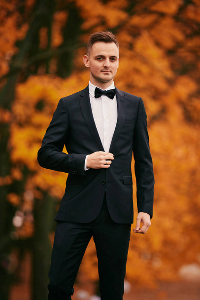 elegante novio joven en un traje negro en el parque de otoño en el día de la boda. - Foto, Imagen