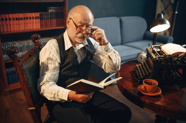 Escritor idoso lê seu trabalho na máquina de escrever vintage em home office. Homem velho em óculos escreve romance de literatura no quarto com fumaça, inspiração
 - Foto, Imagem
