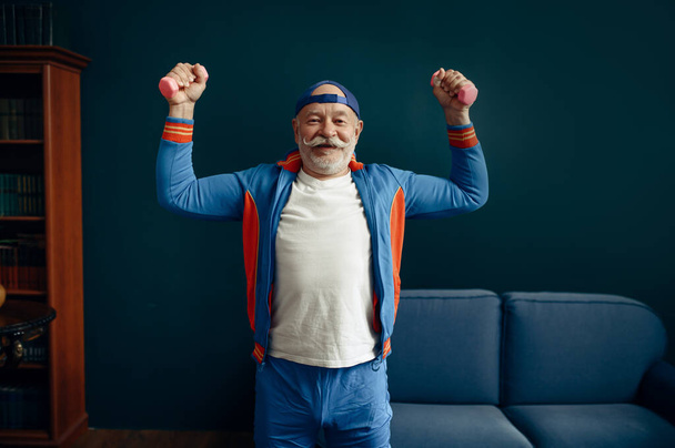 Elderly sportsman in uniform doing exercise with dumbbells at home. Adult male person on fitness training indoors, healthy lifestyle - Foto, immagini