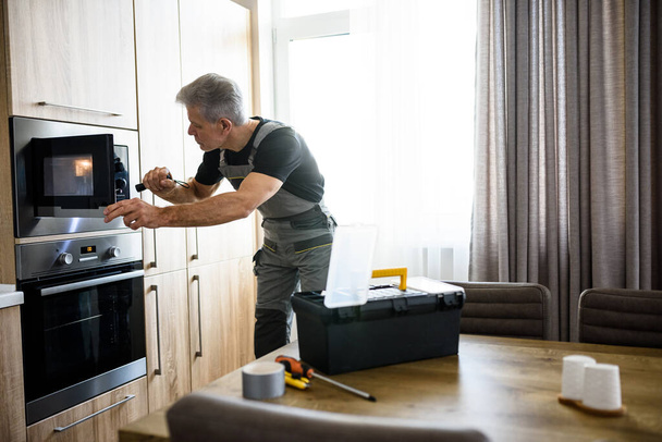 Aged repairman in uniform working, examining broken microwave in the kitchen using flashlight. Repair service concept - Foto, immagini