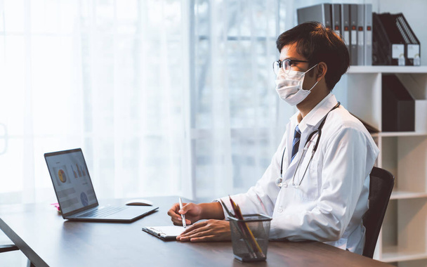 El médico que usa máscara está sentado en un escritorio, que está investigando cómo tratar la condición del paciente.. - Foto, imagen