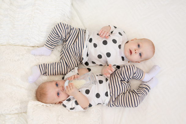 dois gêmeos irmão e irmã 8 meses de idade deitam-se na cama de pijama e bebem leite de uma mamadeira, conceito de comida para bebês, vista superior, conceito de amizade, lugar para texto - Foto, Imagem