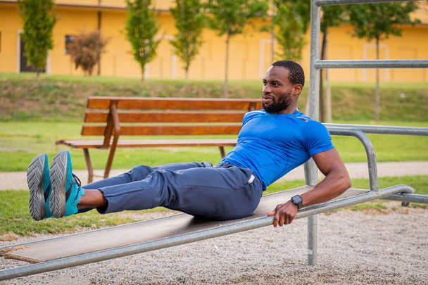 Fitness afro americký chlapec dělá situps venkovní - Fešák africký muž dělá strečink cvičení v open air tělocvičně v parku. Břišní cvičení na lavičce, trénink křupání v City Parku - Fotografie, Obrázek
